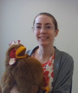 Portrait of Alicia wearing a gray sweater and red adn white top holding a brown chicken puppet