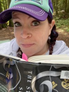 Portrait of BC SRC Co-Chair, Kate Wood, wearing a purple and teal baseball cap and holding a library book.
