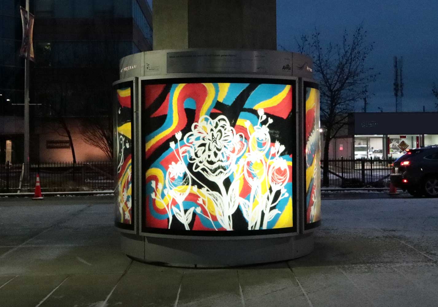 A photograph featuring one of Jeni Chen's public art pieces. It is a white flowers illuminated with a colourful background around a large post.