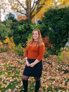 Photograph of BC SRC Content Creator, Erin Crowley, standing outside with fall foliage outside. She is wearing a rust coloured sweater and black skirt.